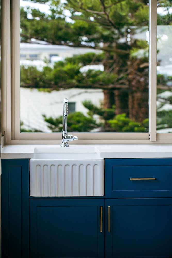 A fluted white butler's sink in navy cabinetry beneath a window of Norfolk pines