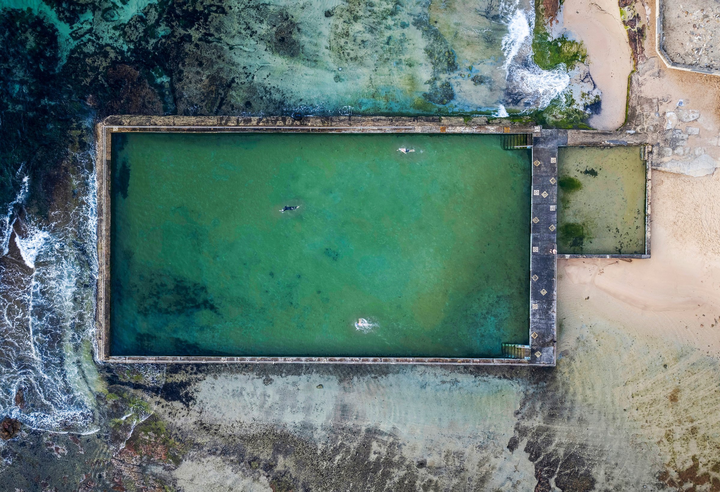Everybody In — Aerial view of swimmers in a rectangular ocean rock pool beside the surf