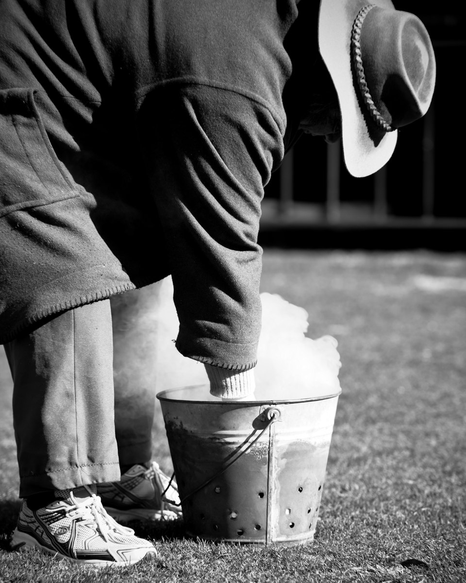 Gather — Black-and-white photograph of a figure in a hat tending a smoking metal bucket