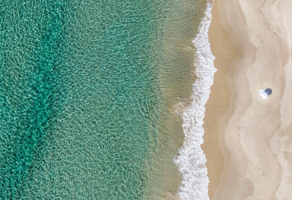 Sundays — Aerial view of a single beach umbrella on pale sand beside the shoreline and clear green water